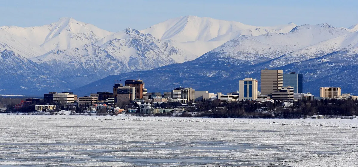 Downtown Anchorage's towers in front of the snow-covered Chugach Mountains, with ice on the water in the foreground
