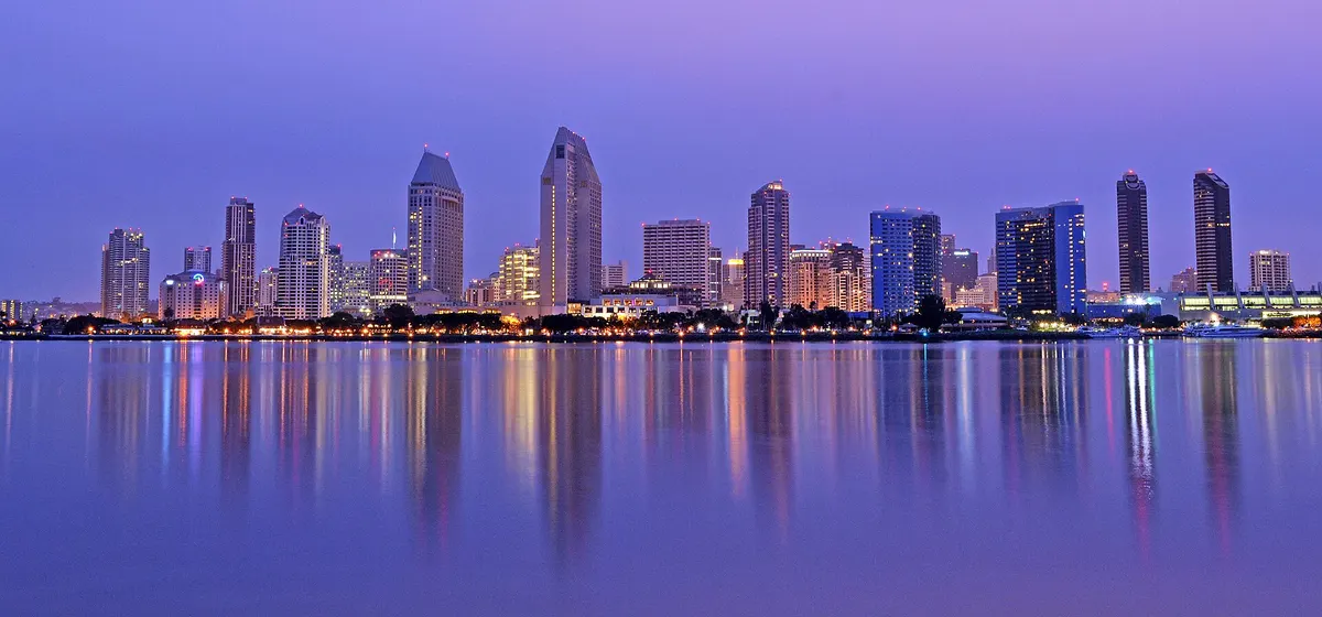 Downtown San Diego's towers at dawn, lit windows reflected in the still water of the bay