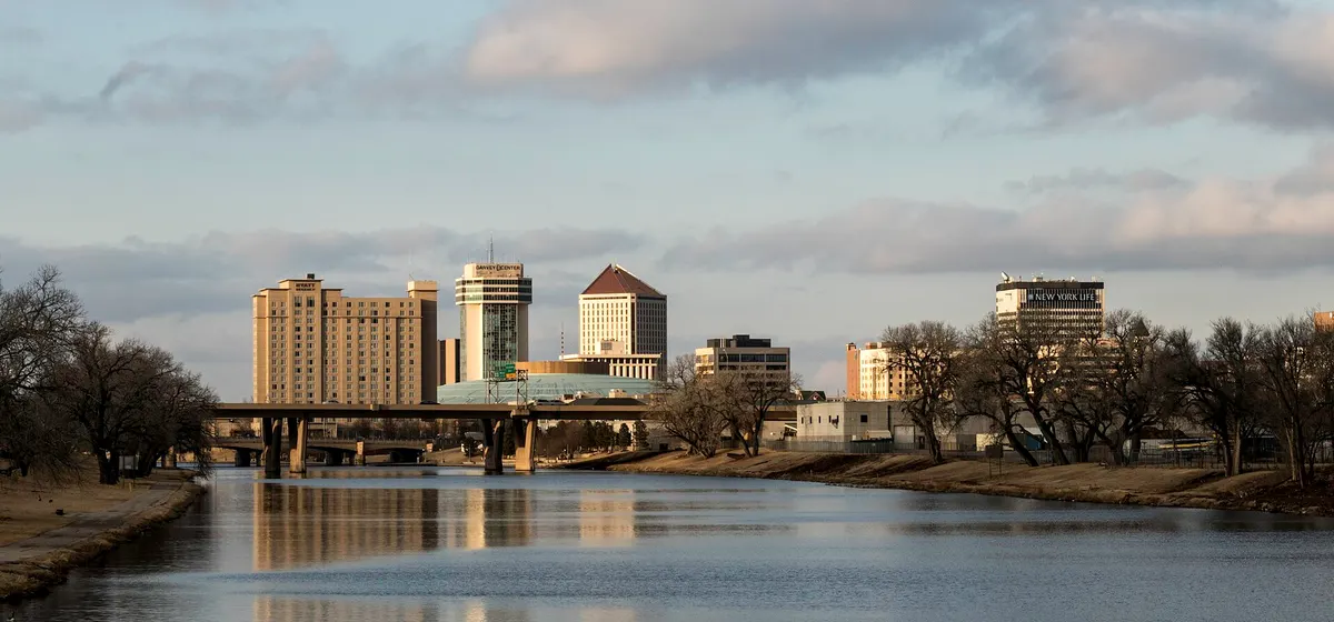 Downtown Wichita's towers beyond the still water of the Arkansas River and a low bridge, under a soft evening sky