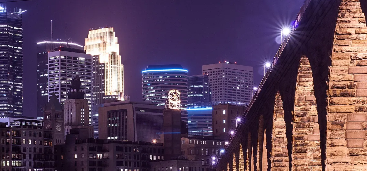 Downtown Minneapolis towers lit at night beside the floodlit arches of the Stone Arch Bridge