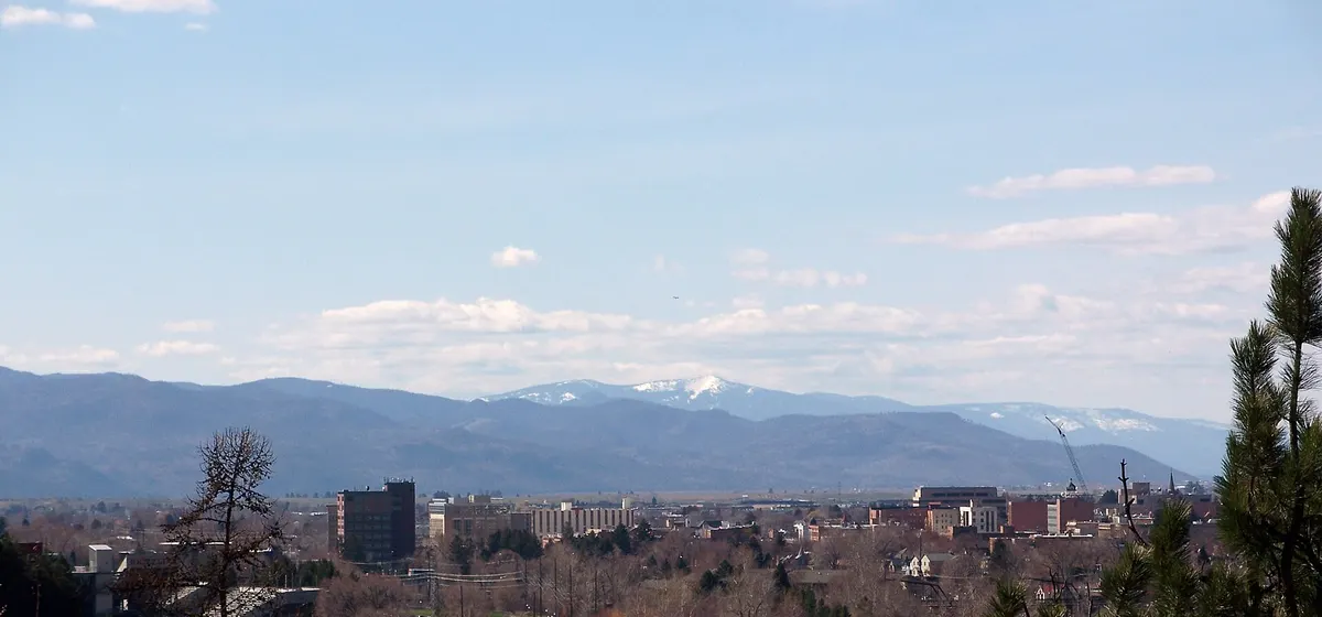 Missoula's low downtown buildings beside a river lined with bare trees, with snow-capped mountains on the horizon