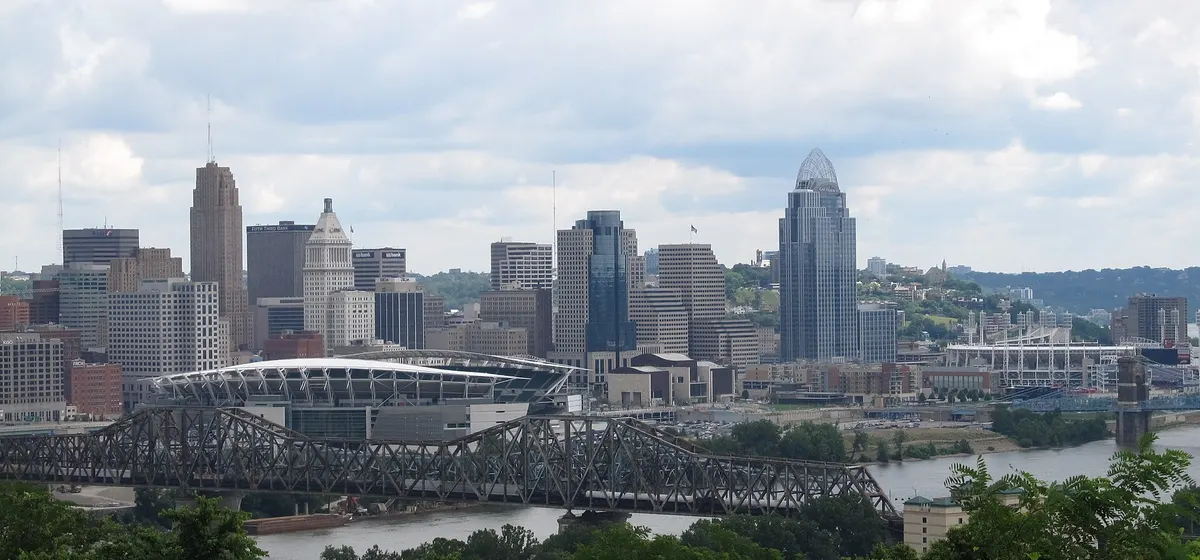 Downtown Cincinnati's towers and riverfront stadium beyond a steel truss bridge over the Ohio River, seen from a wooded hill