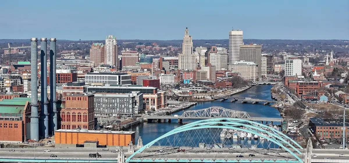 Downtown Providence's towers beyond the Providence River, with a blue arch bridge and brick smokestacks in front