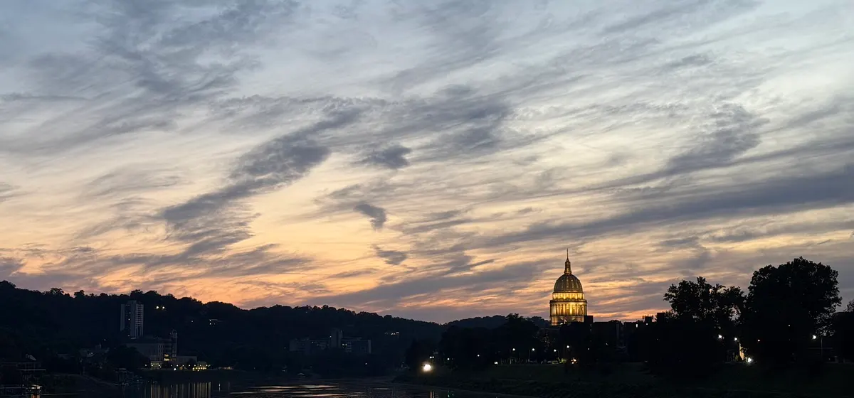 The lit gold dome of the West Virginia State Capitol at twilight, beside the Kanawha River under a streaked evening sky
